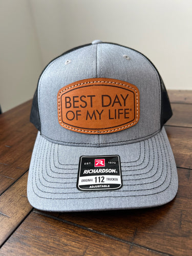 Gray cap with leather patch 'Best Day of My Life' on a wooden surface
